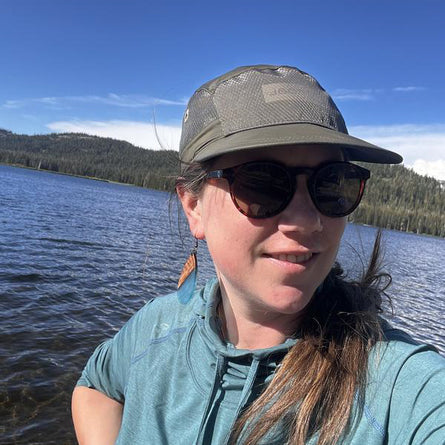 Person wearing sunglasses and a cap by a lake with mountains in the background