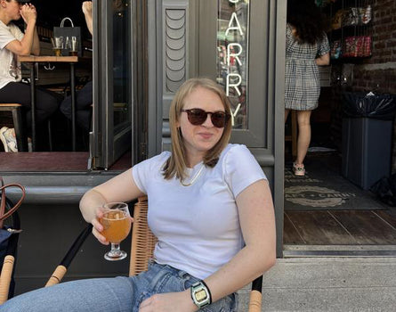 Woman sitting outside a cafe holding a drink, wearing sunglasses and a white shirt.