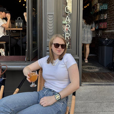 Woman sitting outside a cafe holding a drink, wearing sunglasses and a white shirt.