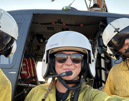 Person in a helicopter cockpit wearing a helmet and sunglasses, with another person partially visible on the right.