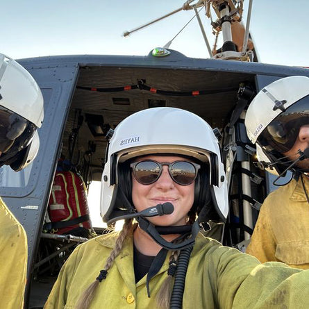 Person in a helicopter cockpit wearing a helmet and sunglasses, with another person partially visible on the right.