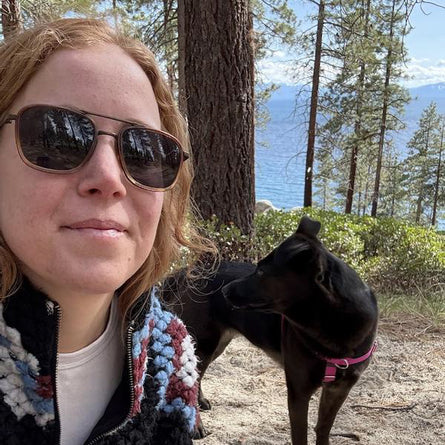 Woman wearing sunglasses with a dog on a trail in a forested area.