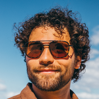 A close-up portrait of a man with curly brown hair and a beard wearing aviator sunglasses with tortoiseshell frames and amber-brown lenses, set against a bright blue sky.