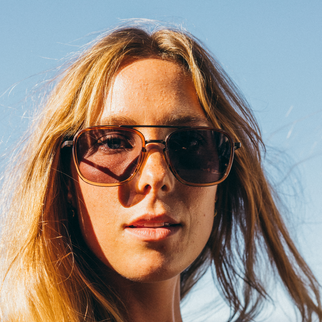 A close-up portrait of a woman with long blonde hair wearing aviator-style sunglasses with dark brown lenses and a thin gold-toned metal frame, set against a clear blue sky.