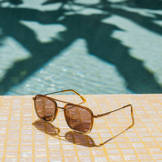 A pair of aviator-style sunglasses with brown tinted lenses and gold metal frames rests on a tiled poolside surface with a swimming pool and leaf shadows in the background.