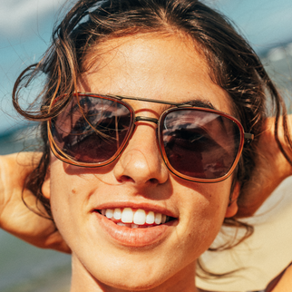 A close-up lifestyle shot of a smiling woman wearing aviator-style sunglasses with brown frames and dark lenses, set against a bright outdoor background.
