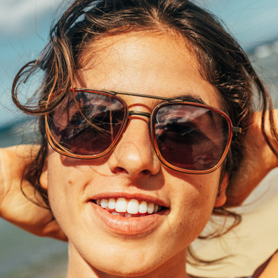 A close-up lifestyle shot of a smiling woman wearing aviator-style sunglasses with brown frames and dark lenses, set against a bright outdoor background.