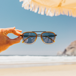 A hand holds a pair of square aviator-style sunglasses with a gold frame and dark lenses against a blurred beach background with ocean, sand, and a sun umbrella.