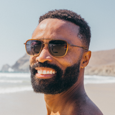 A close-up portrait of a smiling man with a beard wearing aviator sunglasses with gold frames and dark lenses, standing on a sandy beach with ocean and hills in the background.