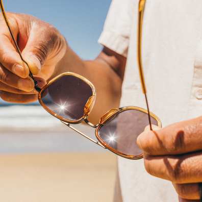 Close-up of a person holding a pair of gold-framed sunglasses with dark lenses, set against a sunny beach background with sand and ocean visible.