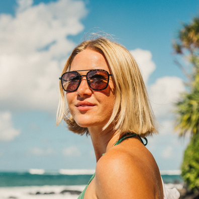 A blonde woman wearing dark-framed sunglasses with mirrored lenses, photographed outdoors on a sunny beach with the ocean and sky in the background.