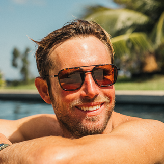 A smiling man with a beard and wet hair relaxes by a pool wearing dark tortoiseshell sunglasses with orange-tinted lenses, with palm trees and blue sky in the background.