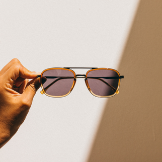 A hand holding a pair of square aviator-style sunglasses with a gold acetate frame and dark lenses, set against a warm beige background with a strong diagonal shadow.