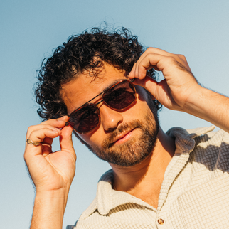 A close-up lifestyle shot of a man with curly dark hair and a beard adjusting aviator sunglasses with dark brown lenses. He is wearing a light beige textured collared shirt against a clear blue sky background.