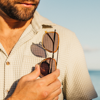 A close-up lifestyle shot of a man holding a pair of tortoiseshell aviator sunglasses in front of his face, wearing a textured beige button-down shirt against a soft-focus beach background.