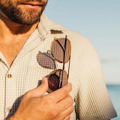A close-up lifestyle shot of a man holding a pair of tortoiseshell aviator sunglasses in front of his face, wearing a textured beige button-down shirt against a soft-focus beach background.