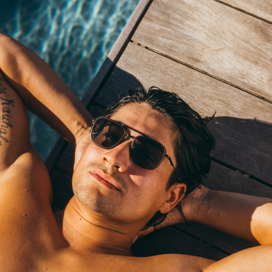 A man wearing dark aviator-style sunglasses relaxes on a wooden poolside deck with blue water in the background.