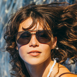 A close-up portrait of a woman with windblown brown hair wearing dark aviator sunglasses, a white beaded necklace, and a silver stud earring against a bright outdoor background.