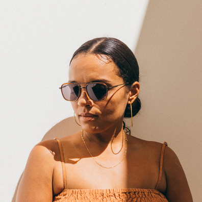 A woman with her hair pulled back wears aviator-style sunglasses with dark lenses and gold-toned frames, along with gold hoop earrings and layered necklaces, against a warm beige wall with strong sunlight and shadows.