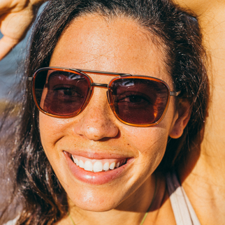 A close-up lifestyle shot of a woman wearing aviator-style sunglasses with tortoiseshell frames and dark brown lenses, smiling outdoors in bright sunlight.