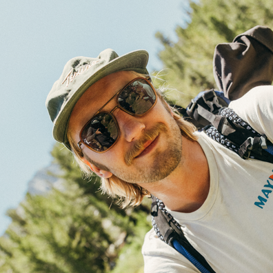 A man outdoors wearing a green cap, sunglasses, a white t-shirt, and a backpack, photographed in a sunny natural setting with trees and blue sky in the background.