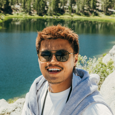 A smiling man outdoors in front of a lake and trees, wearing aviator sunglasses, a white T-shirt, and a light blue zip hoodie. The background shows a natural mountain lakeside setting with rocks and greenery.