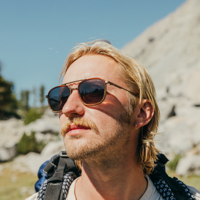 A close-up outdoor portrait of a man wearing aviator-style sunglasses with dark lenses and a gold-tone frame, set against a rocky mountain landscape under a blue sky.