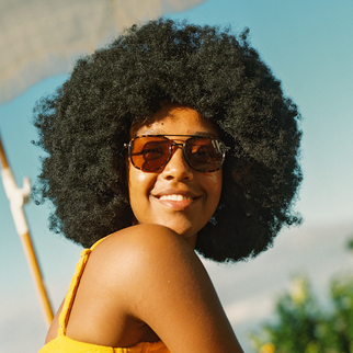 A smiling woman with a large afro hairstyle wearing aviator sunglasses with brown lenses and a yellow top, photographed outdoors against a bright sky.