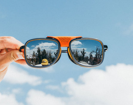 Sunglasses with reflective sky and tree pattern held against a blue sky background