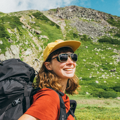A smiling woman hiking on a grassy mountain slope, wearing a yellow cap, dark sunglasses, an orange-red shirt, and carrying a large black backpack.
