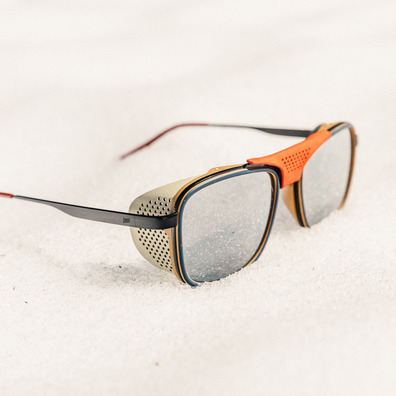 A pair of square shield-style sunglasses with gray tinted lenses, an orange nose bridge, and a beige perforated side shield, resting on a light sandy surface.
