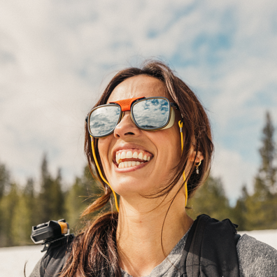 A smiling woman outdoors wearing oversized square sunglasses with a bright orange top frame, black lower frame, and yellow chin straps. The reflective lenses show the sky and trees behind her.