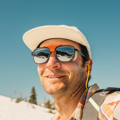 A close-up outdoor portrait of a smiling man wearing a white baseball cap, orange-framed sunglasses, a plaid shirt, and a backpack against a bright blue sky and snowy background.