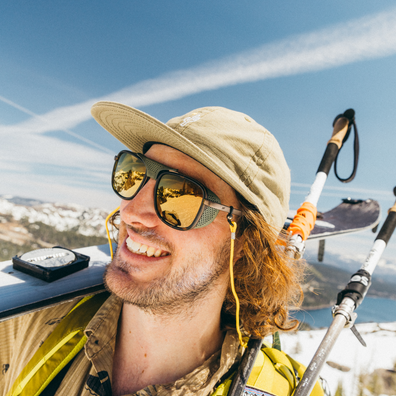 A smiling man outdoors wearing a beige cap, gold-tinted sunglasses, and a yellow-green jacket, with ski poles and a snowboard visible against a blue sky and snowy landscape.
