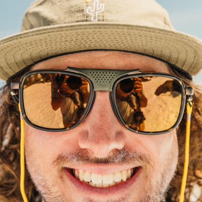 Close-up of a smiling man wearing black sunglasses with reflective gold lenses and a matching gold-tinted strap, along with a beige cap in an outdoor setting.