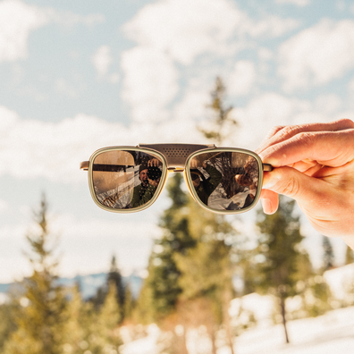 A hand holds a pair of square sunglasses with a gold-toned metal frame and reflective lenses against a blurred outdoor background of snowy trees and blue sky.