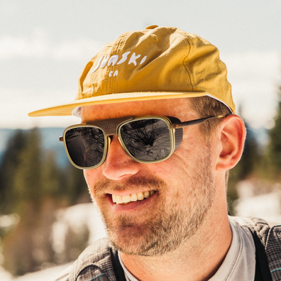 A smiling man outdoors wearing a mustard yellow cap with white 'Jurski' text, dark sunglasses, and a gray plaid shirt, with a blurred snowy mountain background.
