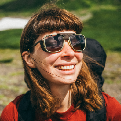 A smiling woman outdoors wearing dark sunglasses with a thick black frame and a black chin strap, set against a blurred green natural background.