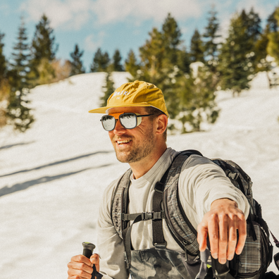 A man hiking in a snowy mountain setting, wearing a yellow cap, sunglasses, a light gray long-sleeve shirt, dark gray bib overalls, and a gray backpack.