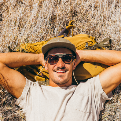 A man lying in dry grass outdoors, wearing a beige t-shirt, a brown cap, and sunglasses, with a large tan hiking backpack behind his head.