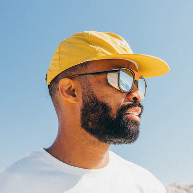 A man in profile wearing a bright yellow baseball cap, black-framed sunglasses with a white lens patch, and a white t-shirt against a clear blue sky.