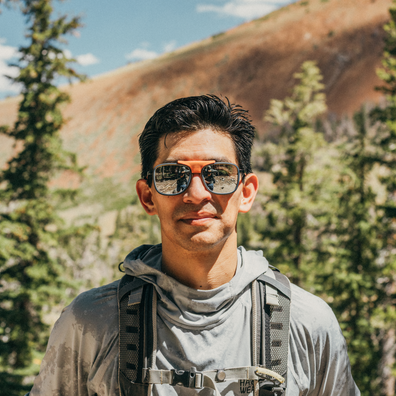 A man wearing orange-framed aviator sunglasses with mirrored lenses, a gray hooded shirt, and a chest strap, photographed outdoors against a mountain and pine tree background.