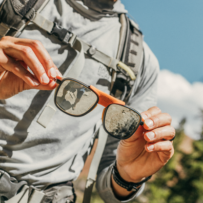 A close-up lifestyle shot of a person outdoors wearing a gray hiking shirt and backpack, holding a pair of sunglasses with orange arms and black frames. The sunglasses have mirrored lenses reflecting the outdoor scenery.