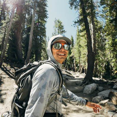 A smiling man wearing a light gray hooded long-sleeve shirt, a gray cap, mirrored sunglasses, and a black hiking backpack, standing on a forest trail surrounded by tall pine trees.