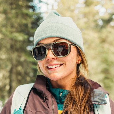 A smiling woman outdoors wearing a light blue knit beanie, dark sunglasses with a gold-tone frame, and a brown and teal jacket. The background is a softly blurred forest.