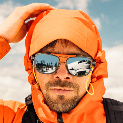 A close-up lifestyle shot of a man wearing an orange hooded jacket and orange-framed sunglasses with a yellow ear loop, set against a blue sky background.