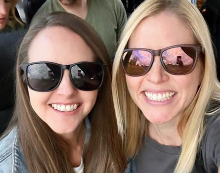 Two women wearing sunglasses with reflective scenery, standing close together.