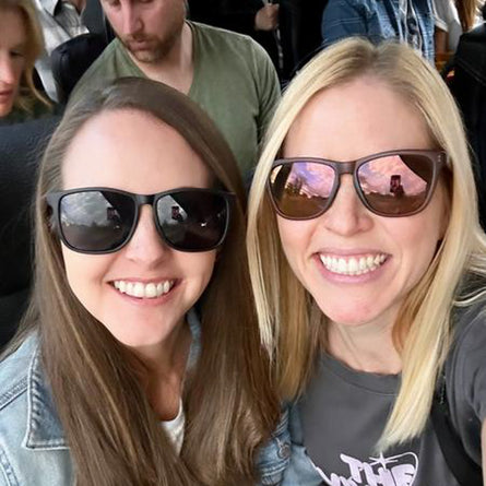 Two women wearing sunglasses with reflective scenery, standing close together.