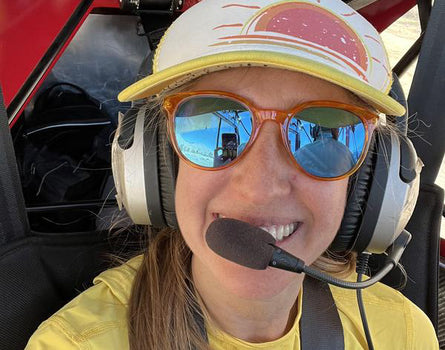 Person wearing a yellow shirt, sunglasses, and a cap with a headset inside a vehicle.