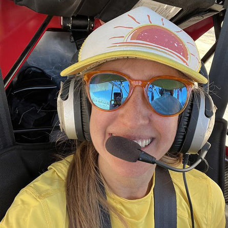 Person wearing a yellow shirt, sunglasses, and a cap with a headset inside a vehicle.